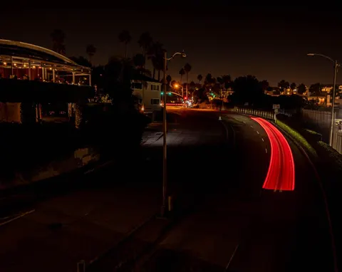 Night long-exposure of red tail-light trails curving past a hillside restaurant patio in Los Angeles, CA
